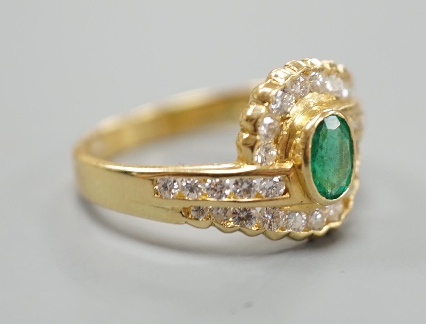 A modern 18ct gold, collet set oval cut emerald and diamond cluster set crossover ring, size N, gross weight 3.5 grams.